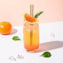 Cocktail in a glass with bagasse straws, garnished with orange and rosemary, surrounded by ice and fruit.