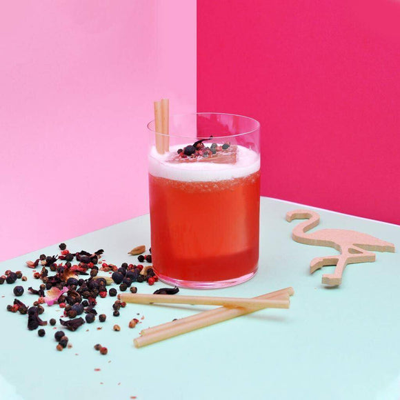Cocktail served with bagasse straws on a colorful background with scattered herbs and a wooden flamingo decoration.