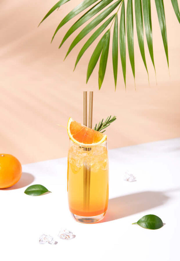 Cocktail glass with a gold straw, orange slice, and palm leaf in a bright setting, showcasing eco-friendly drinking.